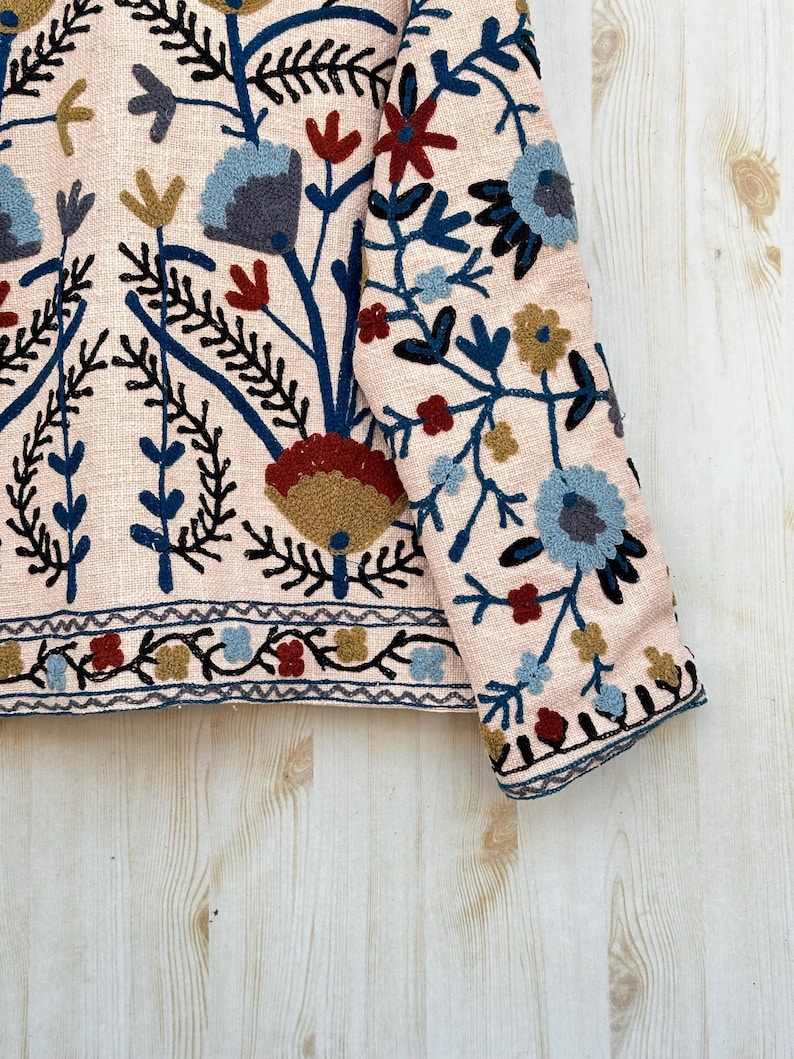Pode incluir: Close-up de um casaco rosa claro com bordados florais intrincados. O design apresenta flores azuis, vermelhas e douradas com caules e folhas pretas. A bainha e os punhos do casaco t&ecirc;m uma borda detalhada.
