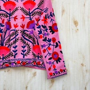 以下が含まれることがあります： 赤、紫、青色の精巧な花の刺繍が施されたピンク色のジャケット。 ジャケットはハイカラーと長袖で、前面と袖には詳細な模様があります。 背景は明るい木目です。