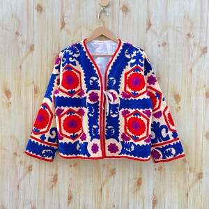 May include: An embroidered jacket in cream, featuring bold blue, red, and purple floral designs. The jacket has a V-neck and a white tie closure. The long sleeves and edges are trimmed in red. The jacket is displayed on a wooden hanger.