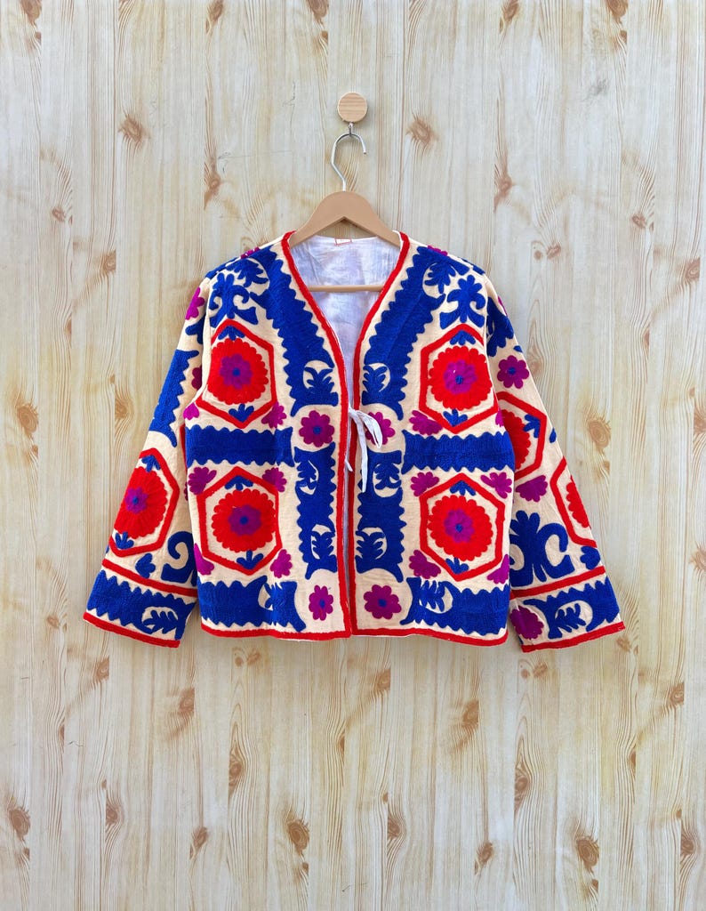 May include: An embroidered jacket in cream, featuring bold blue, red, and purple floral designs. The jacket has a V-neck and a white tie closure. The long sleeves and edges are trimmed in red. The jacket is displayed on a wooden hanger.