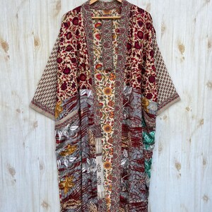 May include: A long, open-front kimono robe with a patchwork design. The robe features a variety of floral patterns in shades of red, beige, gray, and green. The sleeves are wide, and the robe is hanging on a wooden hanger.
