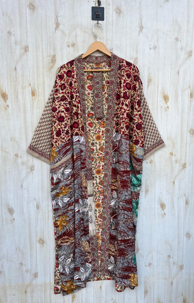 May include: A long, open-front kimono robe with a patchwork design. The robe features a variety of floral patterns in shades of red, beige, gray, and green. The sleeves are wide, and the robe is hanging on a wooden hanger.