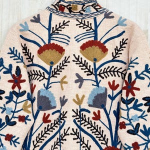 Pode incluir: Casaco rosa claro com bordados florais intrincados. O design apresenta flores e folhas azuis, vermelhas e douradas, com caules e detalhes em preto. O casaco est&aacute; pendurado em um cabide de madeira.