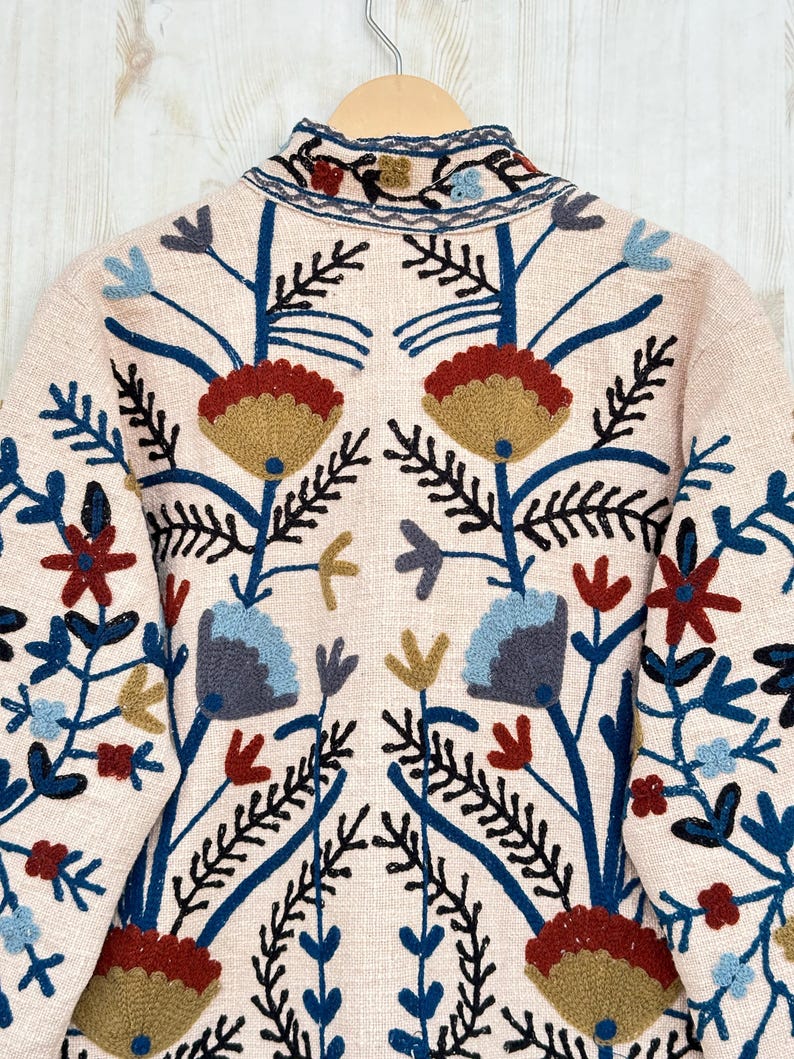 Pode incluir: Casaco rosa claro com bordados florais intrincados. O design apresenta flores e folhas azuis, vermelhas e douradas, com caules e detalhes em preto. O casaco est&aacute; pendurado em um cabide de madeira.