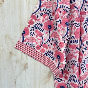 K&ouml;nnte beinhalten: Nahaufnahme eines Stoffes mit floralem Muster. Das Design zeigt rosa Bl&uuml;ten und Bl&auml;tter mit marineblauen Akzenten auf wei&szlig;em Hintergrund. Der Rand des Stoffes hat einen gestreiften Rand in Rosa und Marineblau.