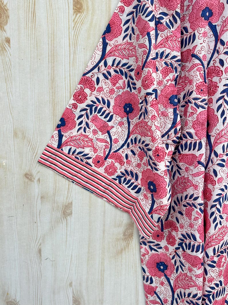 K&ouml;nnte beinhalten: Nahaufnahme eines Stoffes mit floralem Muster. Das Design zeigt rosa Bl&uuml;ten und Bl&auml;tter mit marineblauen Akzenten auf wei&szlig;em Hintergrund. Der Rand des Stoffes hat einen gestreiften Rand in Rosa und Marineblau.