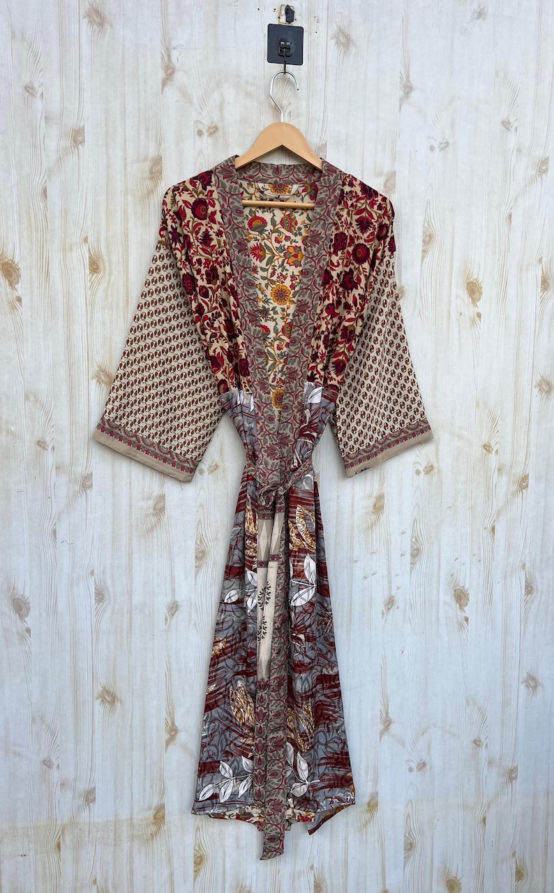 May include: A patterned robe with a floral design in shades of red, beige, and brown hangs on a wooden hanger. The robe features a tie belt and long sleeves with a repeating pattern. The background is a light wood wall.