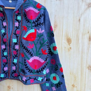 K&ouml;nnte beinhalten: Eine blaue bestickte Jacke mit Blumenmuster. Die Jacke hat einen Stehkragen und lange &Auml;rmel. Die Stickerei ist in den Farben Rosa, Rot, Gr&uuml;n und Schwarz gehalten.
