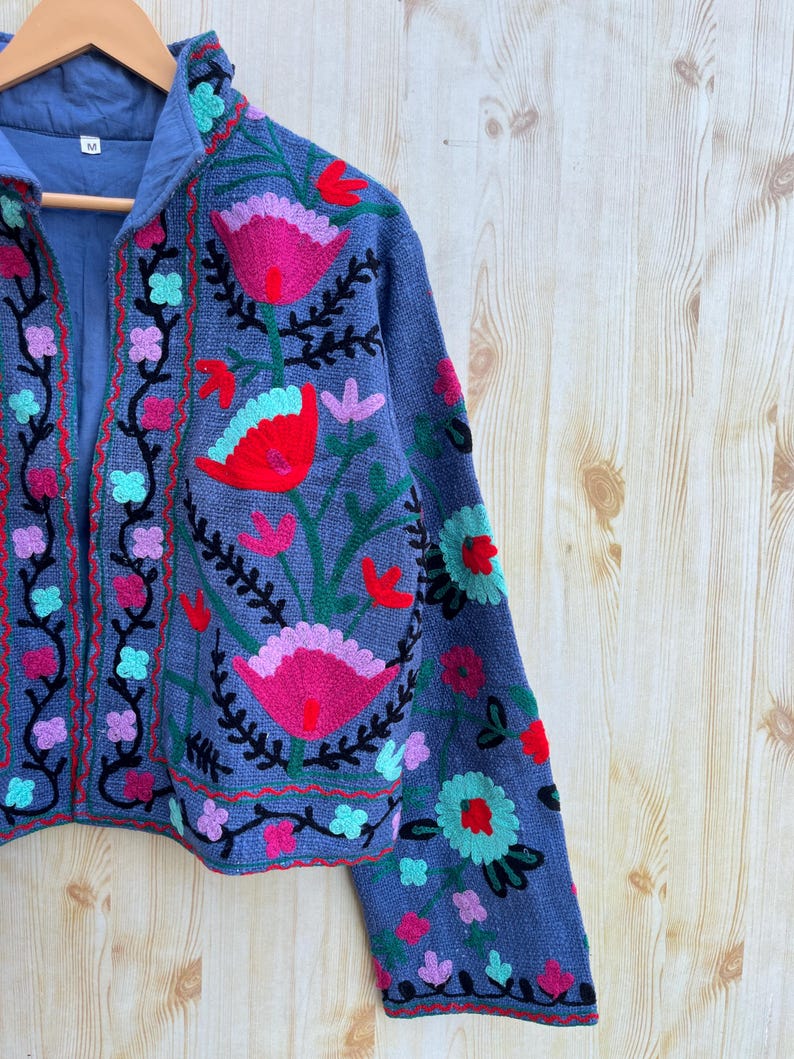K&ouml;nnte beinhalten: Eine blaue bestickte Jacke mit Blumenmuster. Die Jacke hat einen Stehkragen und lange &Auml;rmel. Die Stickerei ist in den Farben Rosa, Rot, Gr&uuml;n und Schwarz gehalten.