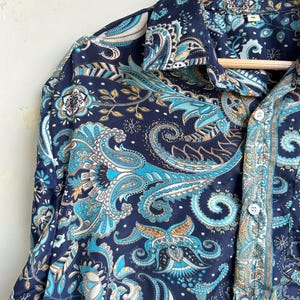 Op de afbeelding: Een marineblauw overhemd met een paisley patroon in tinten van teal, lichtblauw en goud. Het overhemd hangt aan een houten hanger. Het heeft een klassieke kraag en manchetten met knopen. Het overhemd is bedrukt met ingewikkelde bloemmotieven.