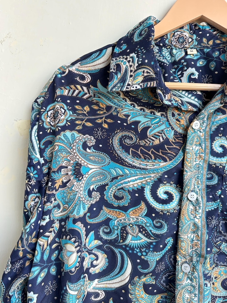 Op de afbeelding: Een marineblauw overhemd met een paisley patroon in tinten van teal, lichtblauw en goud. Het overhemd hangt aan een houten hanger. Het heeft een klassieke kraag en manchetten met knopen. Het overhemd is bedrukt met ingewikkelde bloemmotieven.