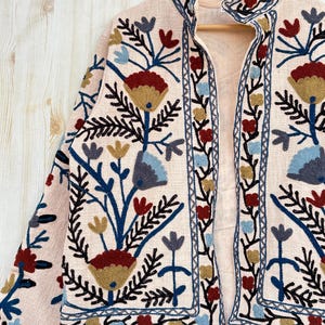 Pode incluir: Casaco bege com bordados florais em vermelho, azul e dourado. O casaco tem gola alta e &eacute; exibido em um cabide de madeira. O bordado inclui flores e folhas estilizadas.