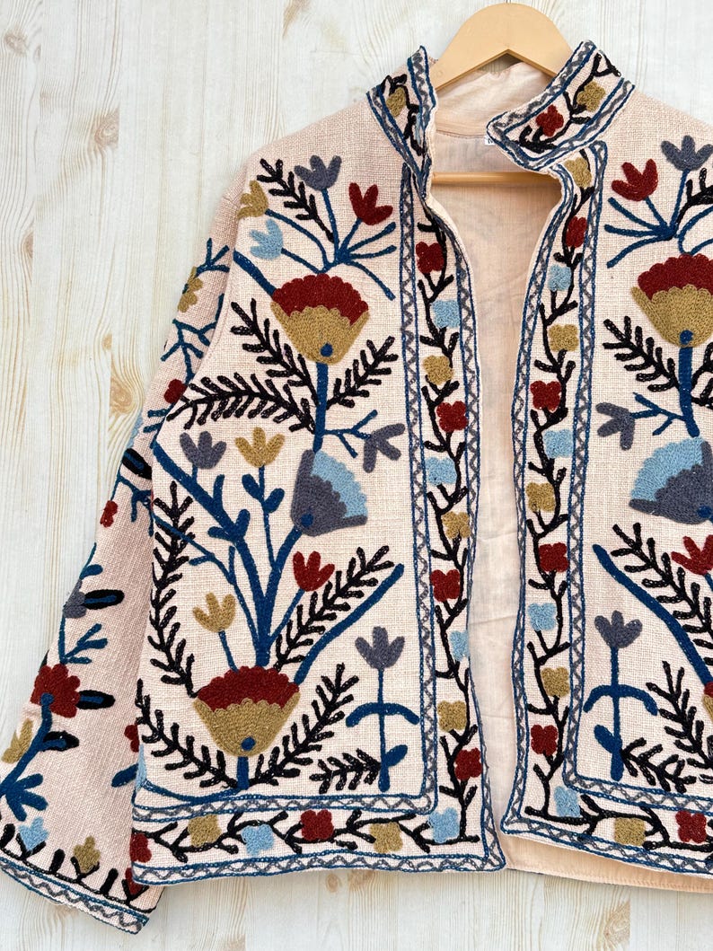 Pode incluir: Casaco bege com bordados florais em vermelho, azul e dourado. O casaco tem gola alta e &eacute; exibido em um cabide de madeira. O bordado inclui flores e folhas estilizadas.