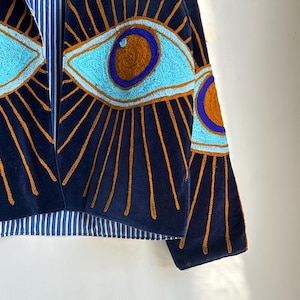 May include: Navy blue velvet jacket with embroidered eye design. The jacket features light blue and brown eye details with gold rays emanating from the eyes. The lining is a blue and white striped fabric.