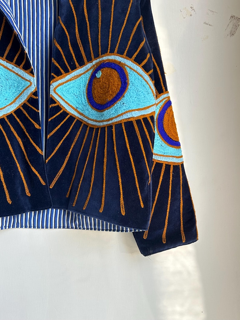 May include: Navy blue velvet jacket with embroidered eye design. The jacket features light blue and brown eye details with gold rays emanating from the eyes. The lining is a blue and white striped fabric.