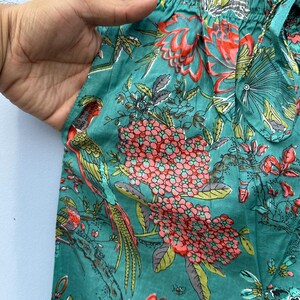 May include: Teal blue cotton fabric with a floral print featuring pink and red flowers, green leaves, and colorful birds. The fabric has a gathered elastic waistband.