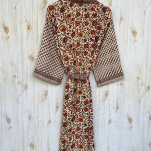 May include: A floral print robe with a cream background, featuring red, orange, and yellow flowers. The sleeves have a repeating geometric pattern in a similar color palette. The robe is hanging on a wooden hanger.