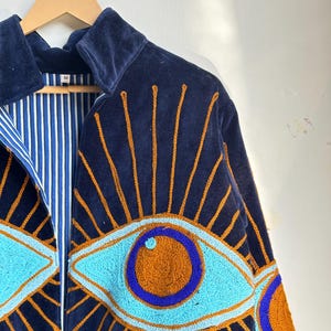 May include: Navy blue velvet jacket with embroidered eye design. The jacket features a light blue and orange eye pattern with gold rays. The interior lining is striped blue and white. The jacket is hanging on a wooden hanger.