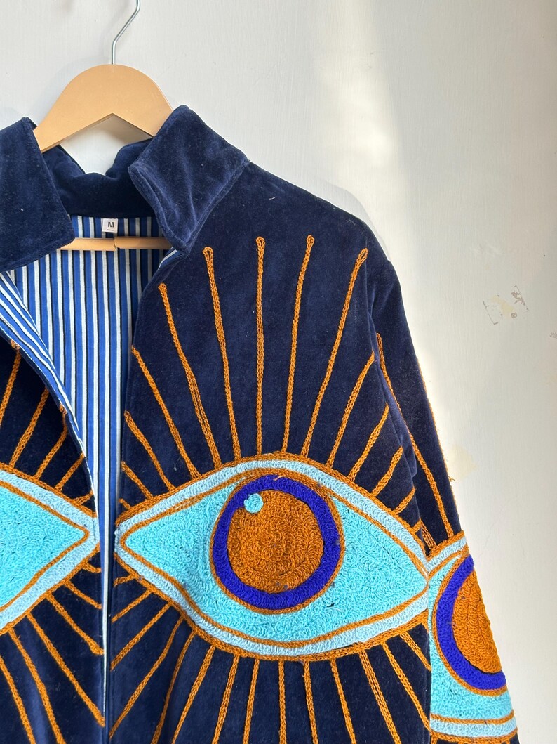 May include: Navy blue velvet jacket with embroidered eye design. The jacket features a light blue and orange eye pattern with gold rays. The interior lining is striped blue and white. The jacket is hanging on a wooden hanger.