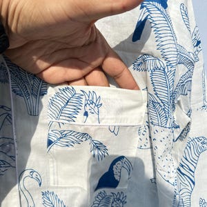 May include: White button-down shirt with a blue and white tropical print featuring toucans, parrots, flamingos, and palm trees. The shirt has a pocket on the left side.