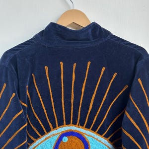 May include: Navy blue velvet jacket with embroidered eye design. The jacket features a high collar and a detailed pattern of turquoise eyes with orange centers and radiating orange lines.