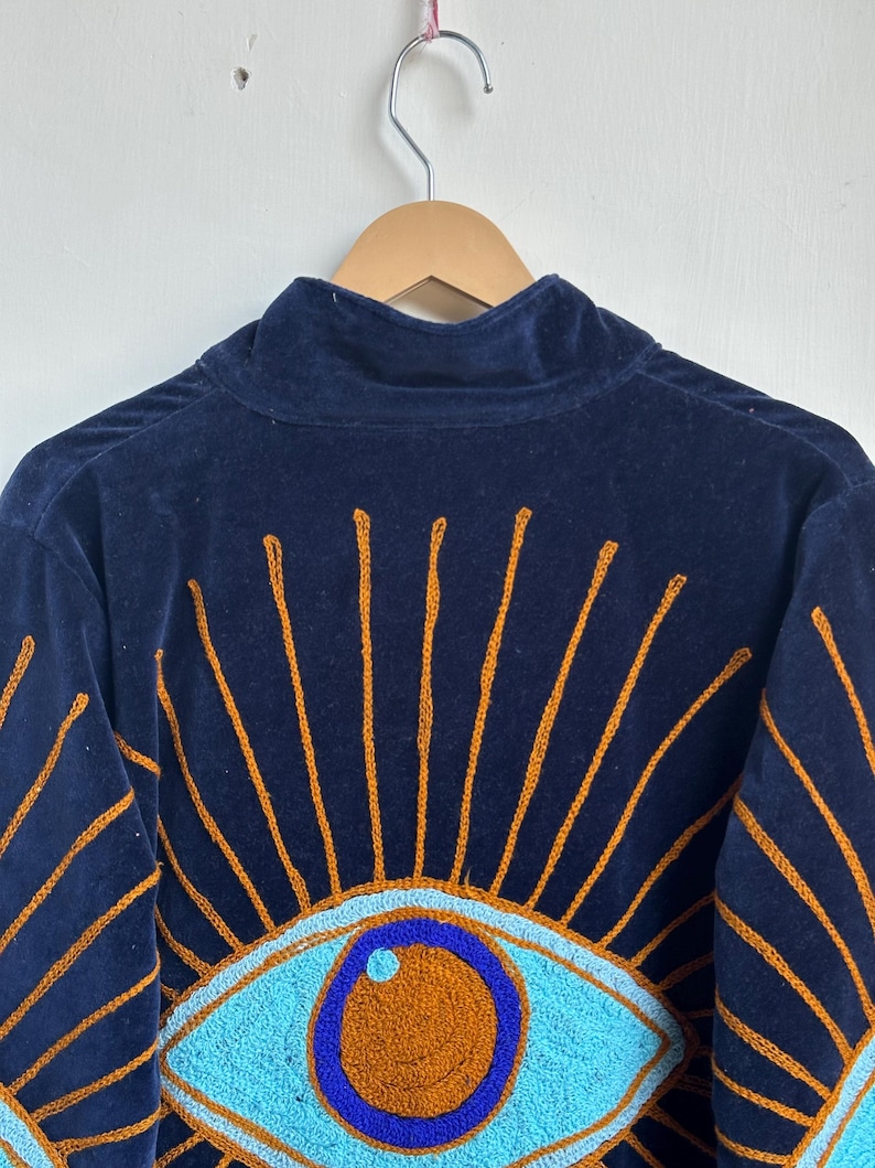 May include: Navy blue velvet jacket with embroidered eye design. The jacket features a high collar and a detailed pattern of turquoise eyes with orange centers and radiating orange lines.