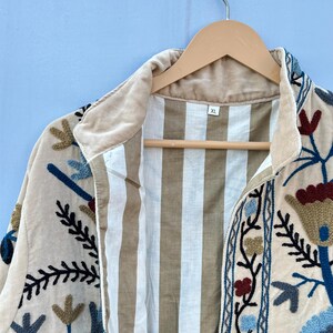 Pu&ograve; includere: Una giacca color crema con ricami floreali in tonalit&agrave; di blu, rosso e marrone. La giacca ha una fodera a righe e un'etichetta di taglia che recita "XL".