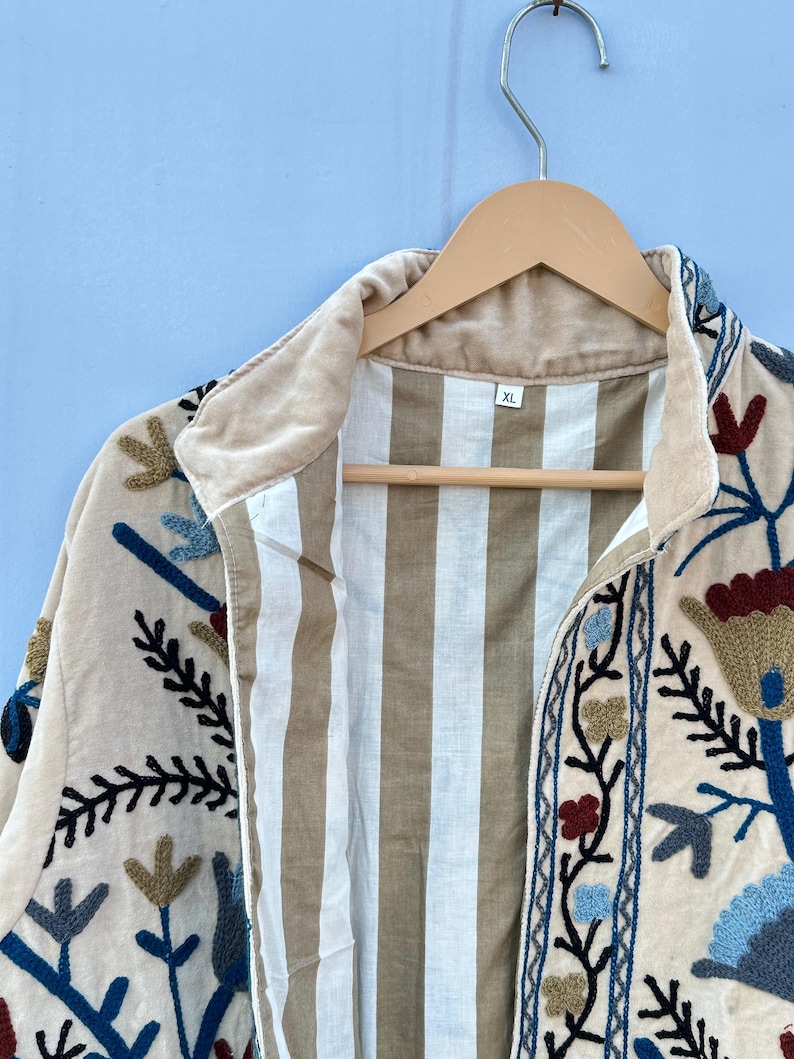 Pu&ograve; includere: Una giacca color crema con ricami floreali in tonalit&agrave; di blu, rosso e marrone. La giacca ha una fodera a righe e un'etichetta di taglia che recita "XL".
