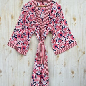 K&ouml;nnte beinhalten: Ein rosa Bademantel mit floralem Muster, gestreiftem Besatz und passendem G&uuml;rtel. Der Bademantel hat einen wei&szlig;en Hintergrund mit rosa Blumen und blauen Akzenten. Der Bademantel h&auml;ngt an einem Holzb&uuml;gel vor einem hellen Holzgrund.