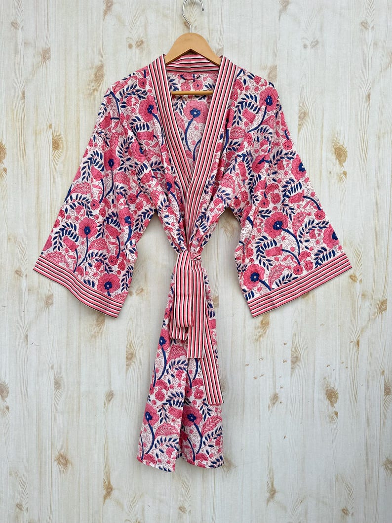 K&ouml;nnte beinhalten: Ein rosa Bademantel mit floralem Muster, gestreiftem Besatz und passendem G&uuml;rtel. Der Bademantel hat einen wei&szlig;en Hintergrund mit rosa Blumen und blauen Akzenten. Der Bademantel h&auml;ngt an einem Holzb&uuml;gel vor einem hellen Holzgrund.