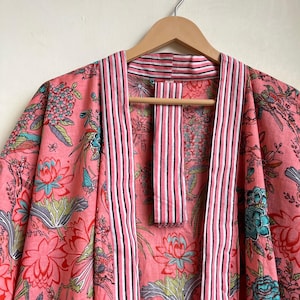 Pu&ograve; includere: Un accappatoio kimono con stampa floreale rosa con finiture a righe. L'accappatoio presenta un motivo vibrante di fiori e uccelli nelle tonalit&agrave; del rosa, del blu e del verde. L'accappatoio &egrave; appeso a un'appendiabiti di legno.
