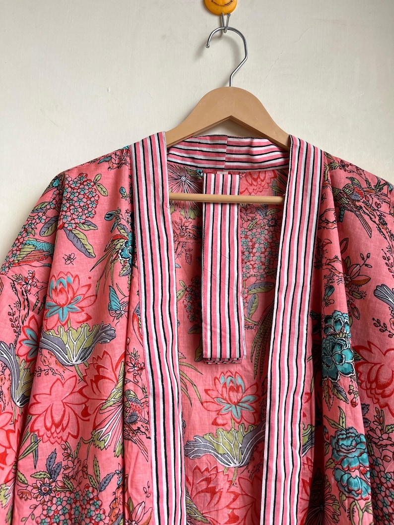 Pu&ograve; includere: Un accappatoio kimono con stampa floreale rosa con finiture a righe. L'accappatoio presenta un motivo vibrante di fiori e uccelli nelle tonalit&agrave; del rosa, del blu e del verde. L'accappatoio &egrave; appeso a un'appendiabiti di legno.