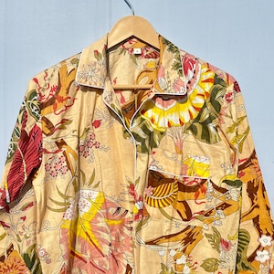 Op de afbeelding: Een beige overhemd met knopen en een bloemenprint in tinten roze, geel, groen en bruin. Het overhemd heeft een witte rand rond de kraag en zakken.