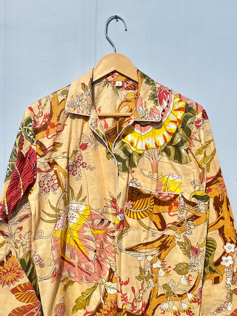 Op de afbeelding: Een beige overhemd met knopen en een bloemenprint in tinten roze, geel, groen en bruin. Het overhemd heeft een witte rand rond de kraag en zakken.