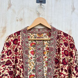 May include: A floral print robe with a cream background and red and burgundy flowers. The robe is hanging on a wooden hanger against a light wood background. The robe has a patterned collar and front panel.
