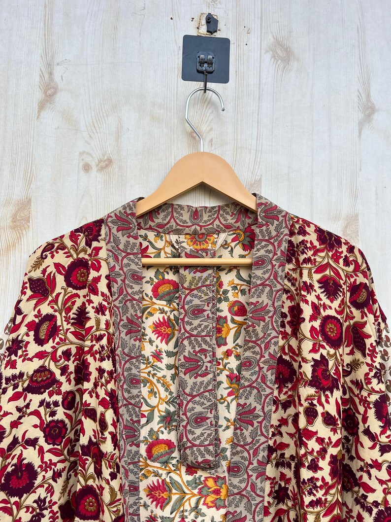 May include: A floral print robe with a cream background and red and burgundy flowers. The robe is hanging on a wooden hanger against a light wood background. The robe has a patterned collar and front panel.