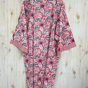K&ouml;nnte beinhalten: Kimono-Robe mit floralem Muster in Rosa, Marineblau und Wei&szlig;. Die Robe zeigt ein sich wiederholendes Muster aus stilisierten Blumen und Bl&auml;ttern. Sie h&auml;ngt an einem Holzb&uuml;gel vor einem hellen Holzhintergrund.