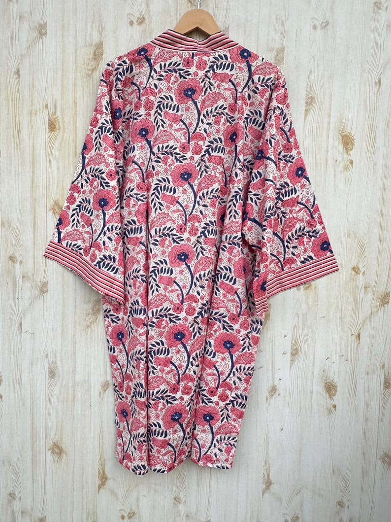 K&ouml;nnte beinhalten: Kimono-Robe mit floralem Muster in Rosa, Marineblau und Wei&szlig;. Die Robe zeigt ein sich wiederholendes Muster aus stilisierten Blumen und Bl&auml;ttern. Sie h&auml;ngt an einem Holzb&uuml;gel vor einem hellen Holzhintergrund.