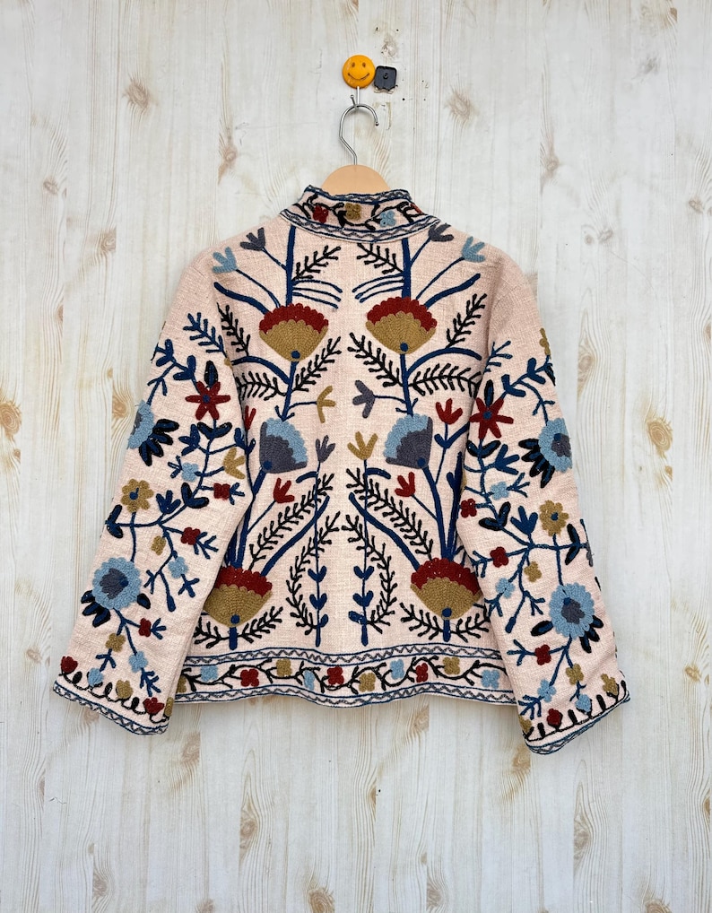 Pode incluir: Casaco rosa claro com bordados florais intrincados em tons de azul, vermelho, amarelo e castanho. O casaco tem gola alta e est&aacute; pendurado em um cabide de madeira contra um fundo de madeira clara. O bordado cria um design sim&eacute;trico.