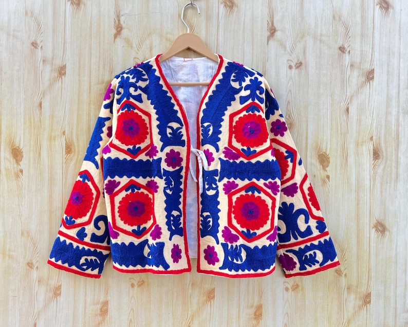 May include: A cream-coloured jacket with detailed embroidery in red, blue, and purple. The jacket has a floral design and a tie closure. It is displayed on a wooden hanger against a light wood backdrop.