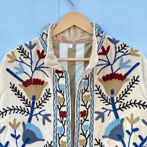 Pu&ograve; includere: Una giacca color crema con un ricamo floreale in tonalit&agrave; di blu, rosso e oro. La giacca ha un colletto e una chiusura con cerniera frontale. L'etichetta della taglia sulla giacca riporta "XL".