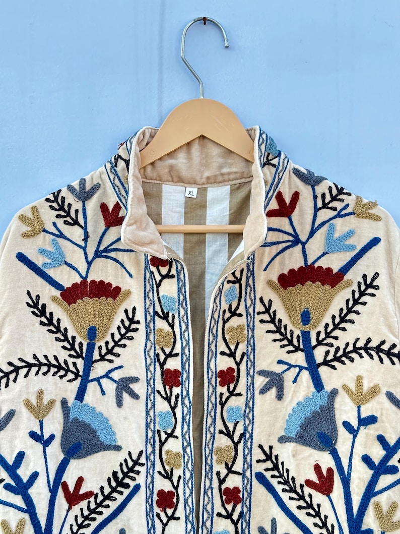Pu&ograve; includere: Una giacca color crema con un ricamo floreale in tonalit&agrave; di blu, rosso e oro. La giacca ha un colletto e una chiusura con cerniera frontale. L'etichetta della taglia sulla giacca riporta "XL".