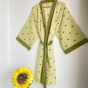 May include: A light green and white striped robe with red heart accents. The robe has olive green trim and a matching belt. The robe is hanging on a wooden hanger against a white wall.