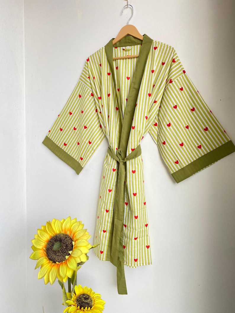 May include: A light green and white striped robe with red heart accents. The robe has olive green trim and a matching belt. The robe is hanging on a wooden hanger against a white wall.