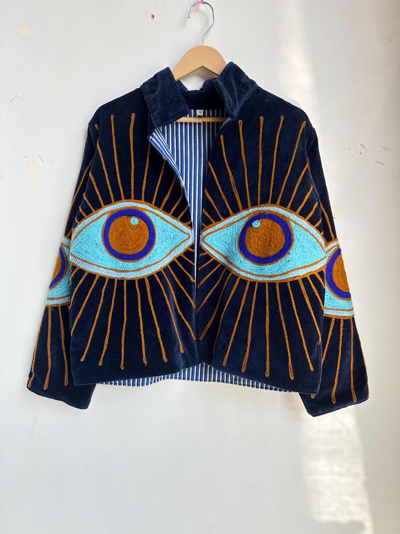 May include: A navy blue velvet jacket with embroidered eye designs. The jacket features light blue and brown eye details with gold sunburst rays. The interior lining has a blue and white striped pattern. The jacket is hanging on a wooden hanger.