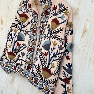 Pode incluir: Casaco rosa claro com bordados florais intrincados em tons de azul, vermelho e dourado. O casaco tem gola alta e &eacute; exibido em um cabide de madeira. O bordado cria um visual texturizado e decorativo.