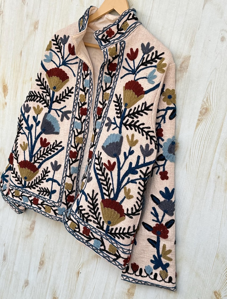 Pode incluir: Casaco rosa claro com bordados florais intrincados em tons de azul, vermelho e dourado. O casaco tem gola alta e &eacute; exibido em um cabide de madeira. O bordado cria um visual texturizado e decorativo.