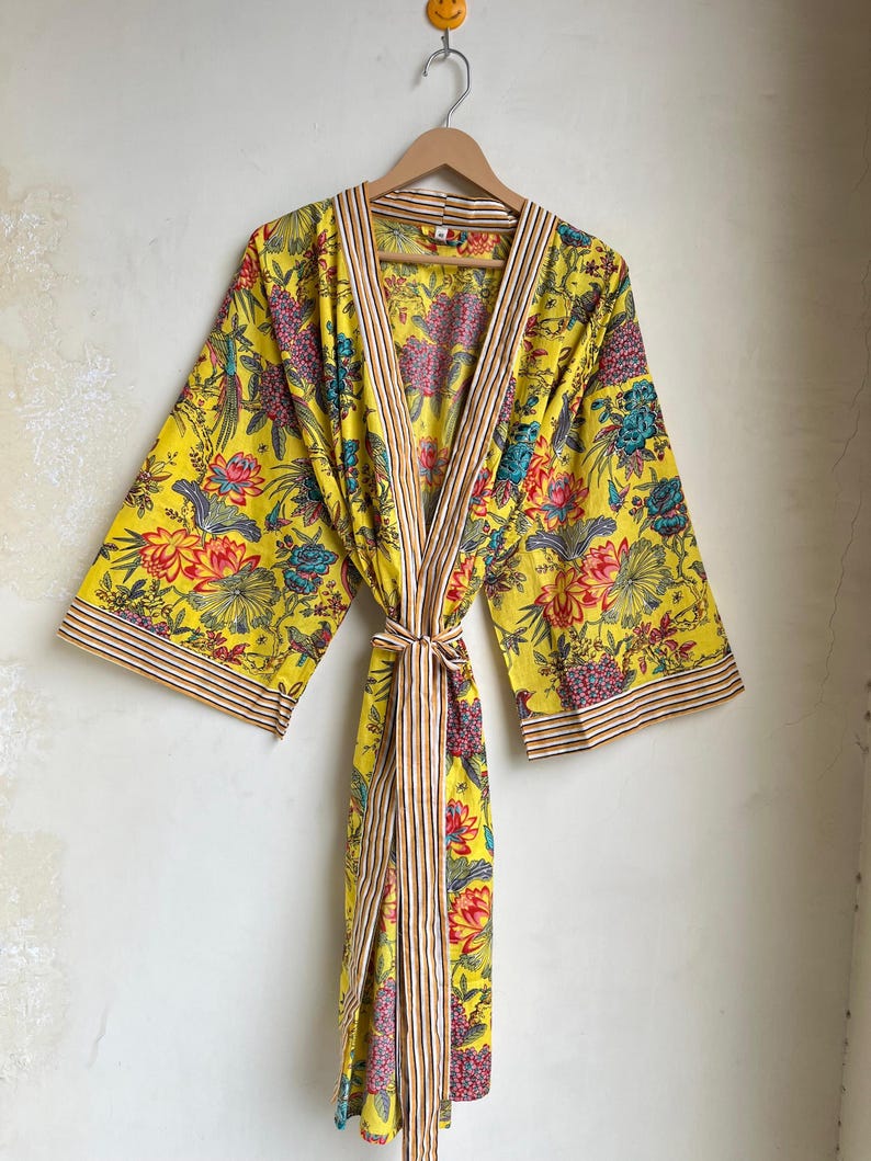 Pu&ograve; includere: Un accappatoio giallo con stampa floreale, finiture a righe e cintura abbinata. L'accappatoio presenta un vivace motivo di fiori e uccelli rossi, blu e rosa. L'accappatoio &egrave; appeso a un'appendiabiti di legno.