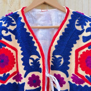 May include: Cream-coloured embroidered jacket with intricate blue and red floral patterns. The jacket features a V-neck design with a red trim and white tie closure. The embroidery includes purple flower accents.