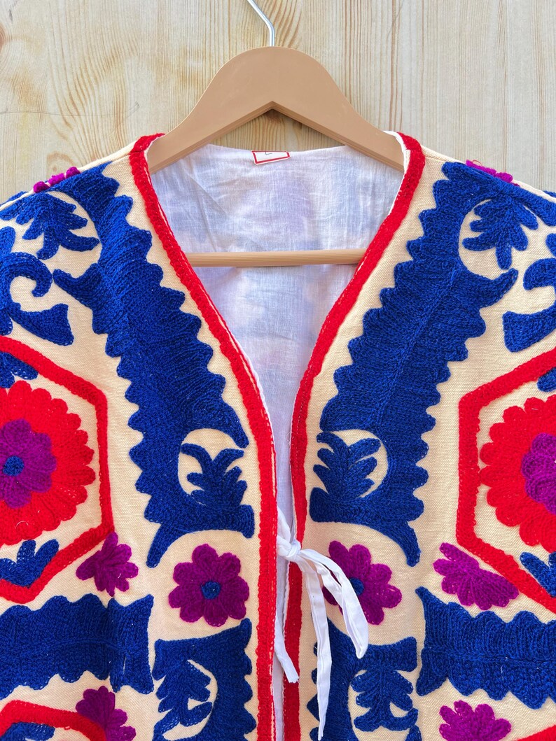 May include: Cream-coloured embroidered jacket with intricate blue and red floral patterns. The jacket features a V-neck design with a red trim and white tie closure. The embroidery includes purple flower accents.